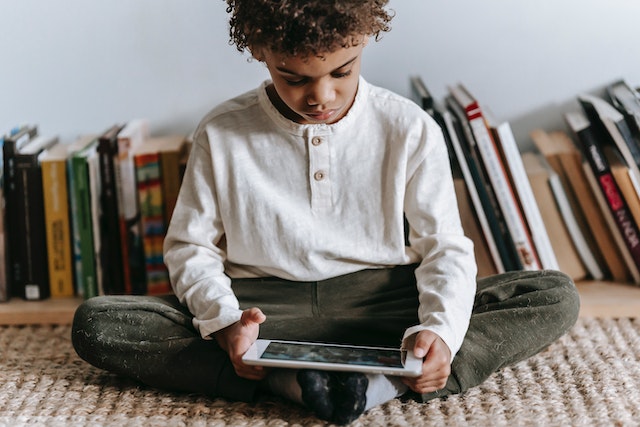 A boy playing on his tablet on the carpet with a bookshelf behind him
