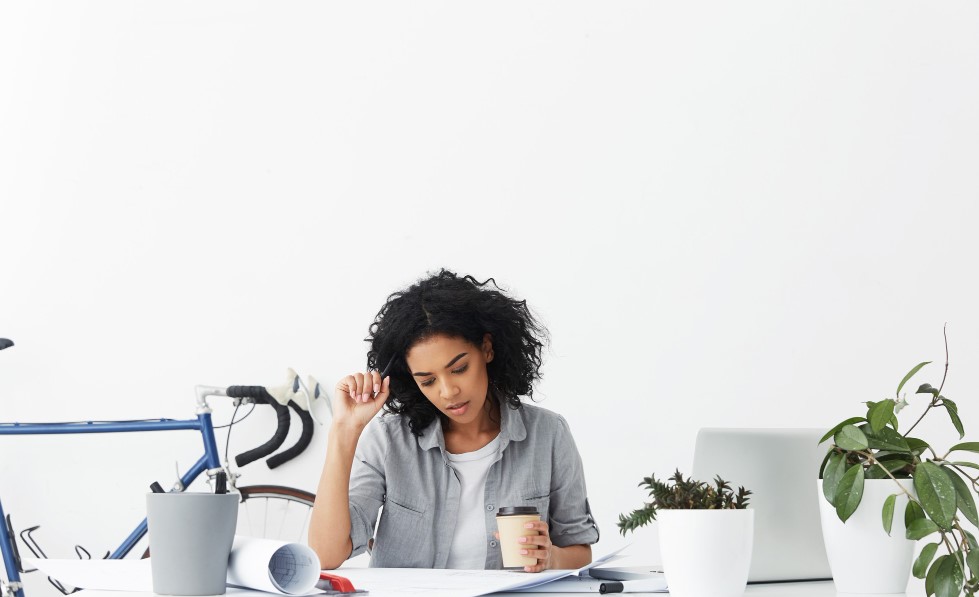 A lady in her home office holding a cup of coffee and a pen to her head trying to be productive.