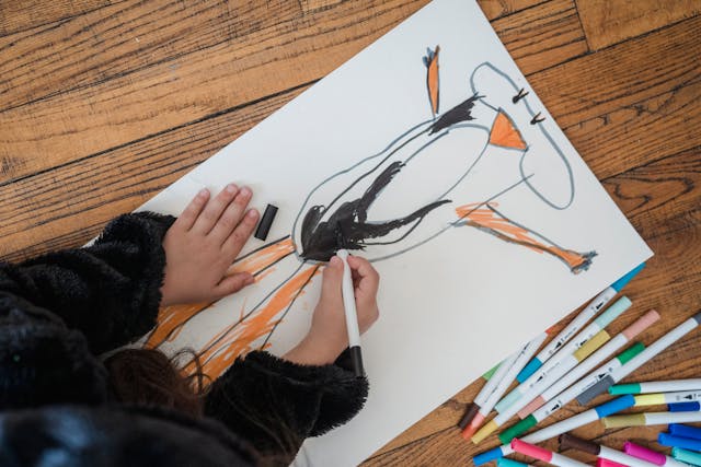 A top view of a child drawing with markers on paper on the wooden floor