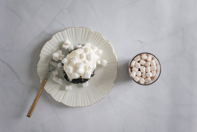 A s’mores cupcake sitting on a plate next to a cup of marshmallows on a marble surface