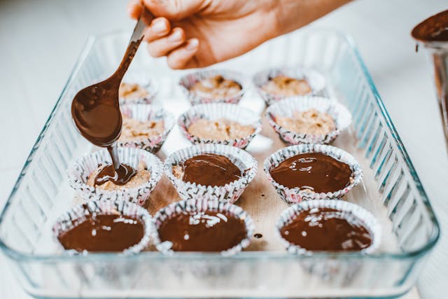 A baker pouring melted chocolate on top of twelve cannoli cupcakes in a baking dish