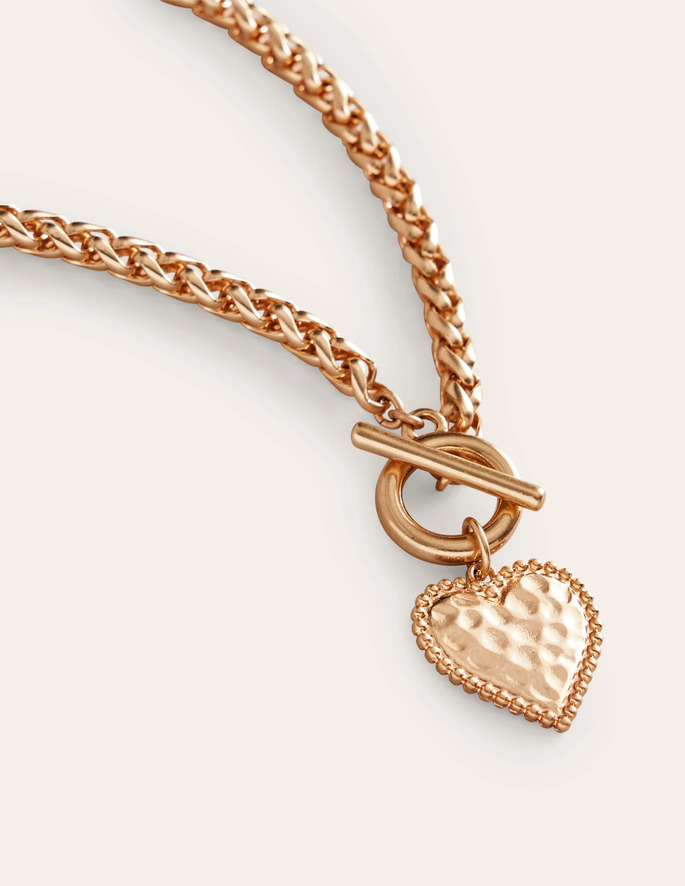 A gold-toned hammered metal heart necklace laying on an off-white background