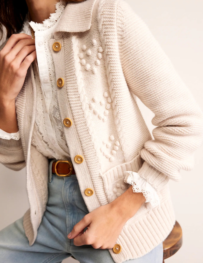 A model sitting on a stool posing in a white lace shirt, blue jeans and a cream cardigan