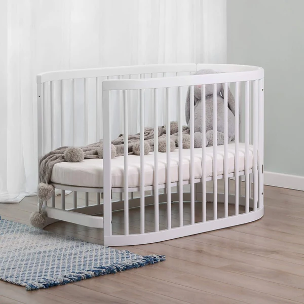 Image: A white oval-shaped cot bed standing on a wooden floor in front of a blue rug A white oval-shaped cot bed standing on a wooden floor in front of a blue rug