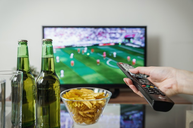 Two beer bottles and chips next to a hand pointing a remote at a flat-screen TV to watch the FA Cup