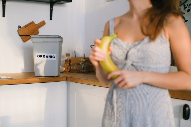 A woman holding a banana standing in front of a counter with a compost bin