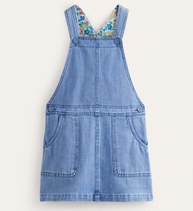 Image: Boden baby girl clothes: A denim dungaree dress with floral lining laying on a beige background Boden baby girl clothes: A denim dungaree dress with floral lining laying on a beige background