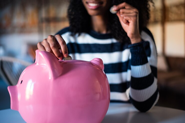Image: Things to do this summer holidays: Pink piggy bank with lady in the background stretching out her hand and putting money in it. Things to do this summer holidays: Pink piggy bank with lady in the background stretching out her hand and putting money in it.