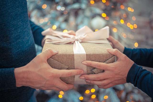 Two people exchanging a wrapped gift with a bow in front of a white fairy tree lit Christmas tree