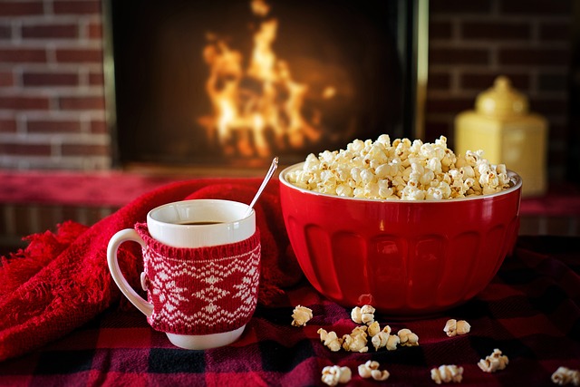 Popcorn and hot chocolate placed on a black and red tablecloth in front of a roaring fireplace
