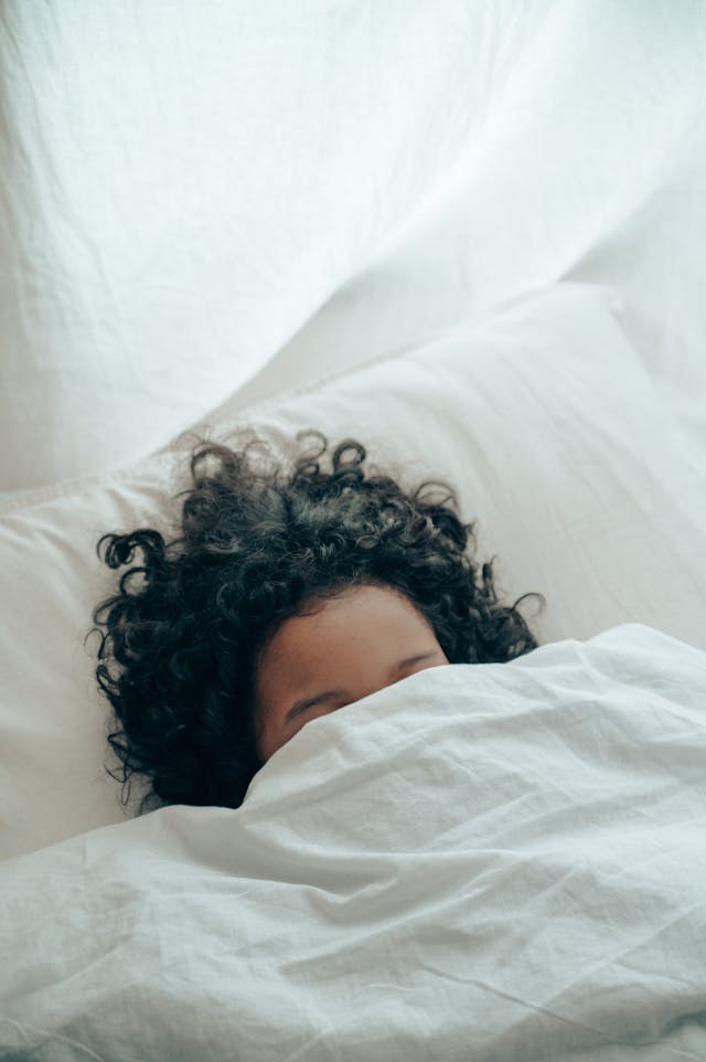 A person with curly hair hiding under a white duvet while lying against a white pillow in bed