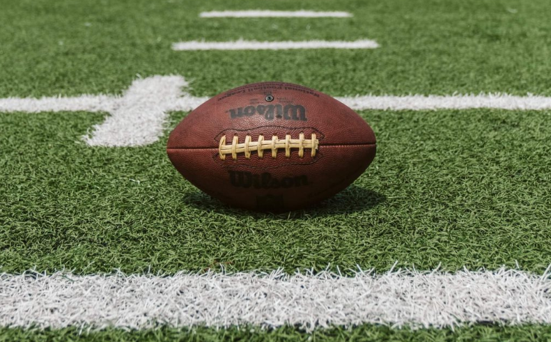 A picture showing a brown Wilson football ball on green turf with white markings