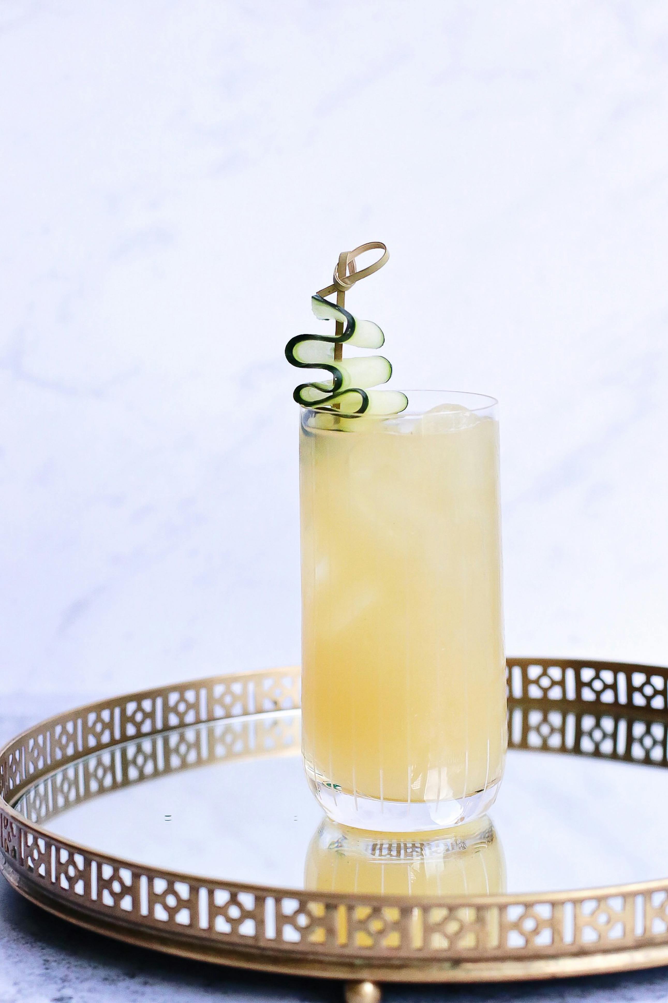 A glass of a ginger drink with zucchini garnish on a glass decorative tray.
