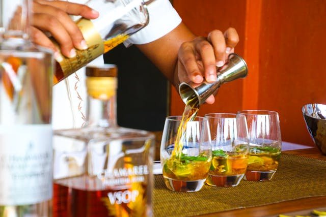 A bartender pouring a shot of alcohol in a glass containing a cocktail from behind a bar.