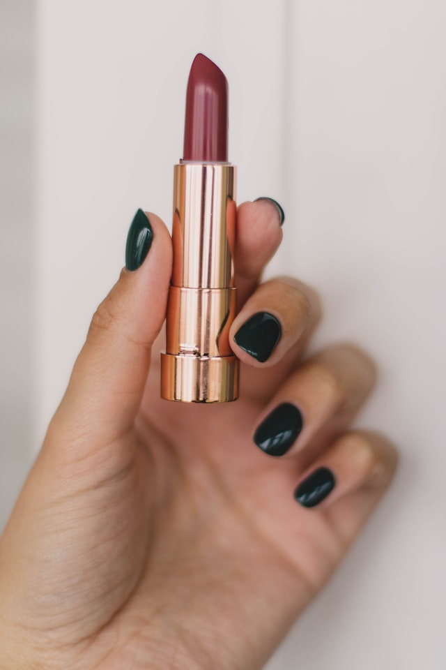 A hand with black painted nails holding a tube of red lipstick.