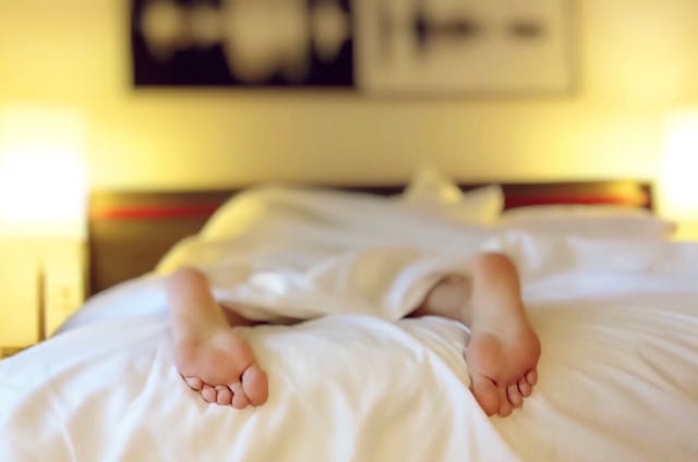 A person lying face down on a white mattress with only their feet being exposed from under the duvet.