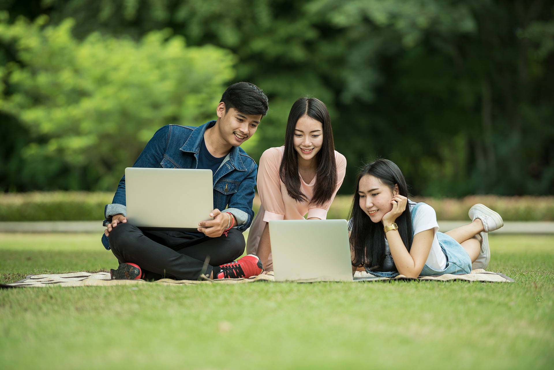 Three students are sitting on the grass looking at two laptops, looking for Windsor Smith promo codes.