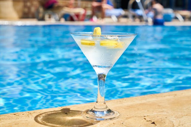 A close-up of a martini cocktail topped with lemon slices sitting on the side of a pool.