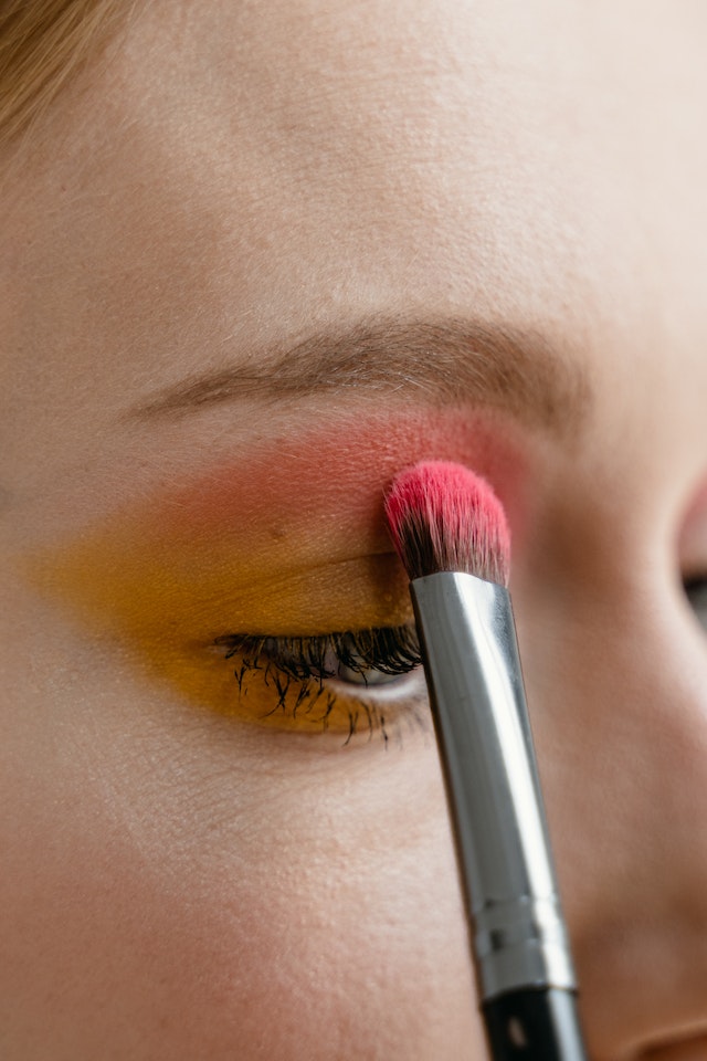 A woman applying pink eye shadow on top of yellow eyeshadow with a brush.