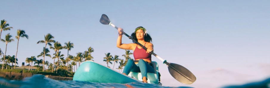 A woman sitting on a blue paddle board while paddling. A clear sky on the background and some trees showing