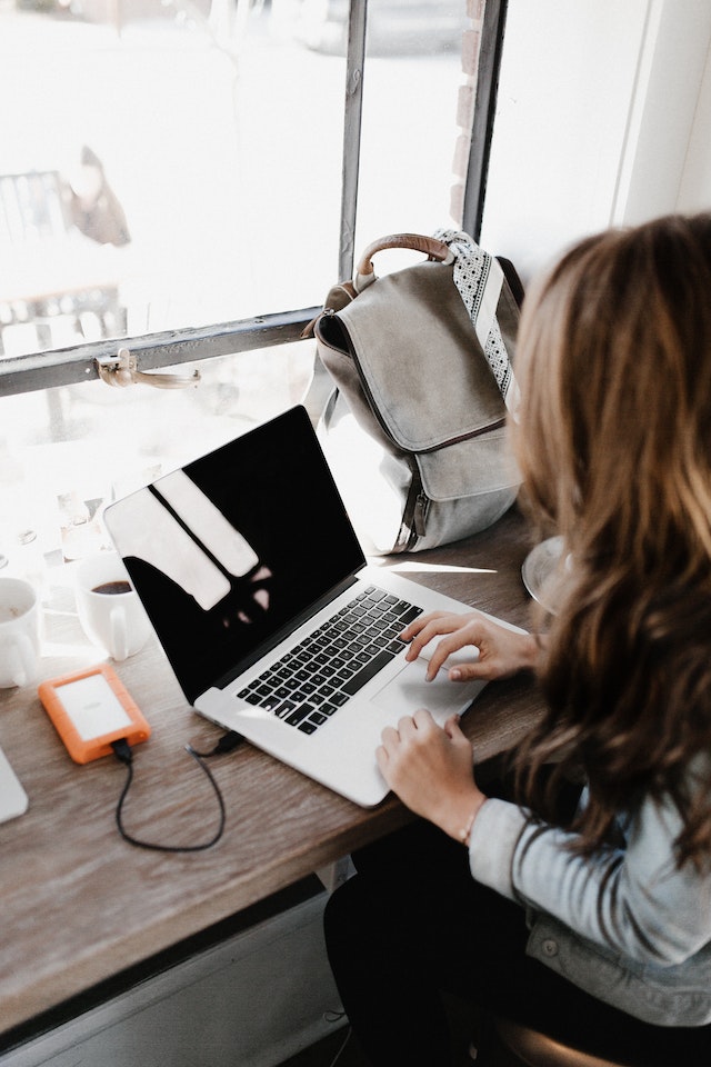 Person typing on a laptop while looking at a Hearst Magazines promo code with a mobile phone, cup and some paper with notes written on sitting on a wooden desk