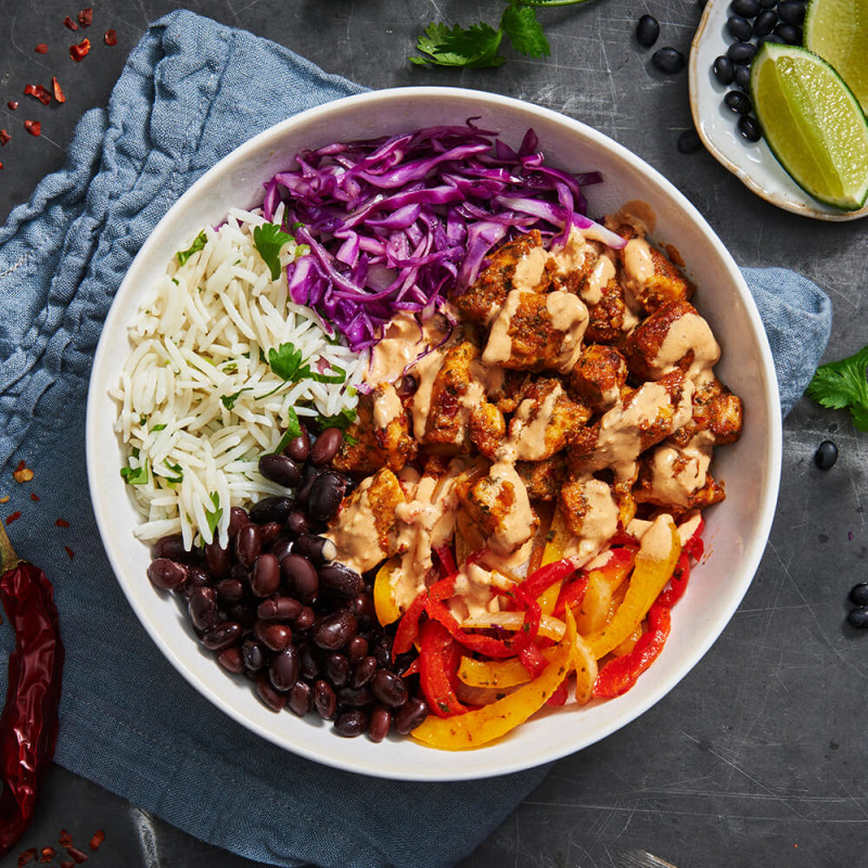 chicken burrito bowl seated on a napkin and surrounded by limes, beans, herbs and chilis