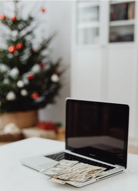 Christmas tree blurred out in the background with a laptop and money on it encapsulating the idea of saving money during Christmas shopping