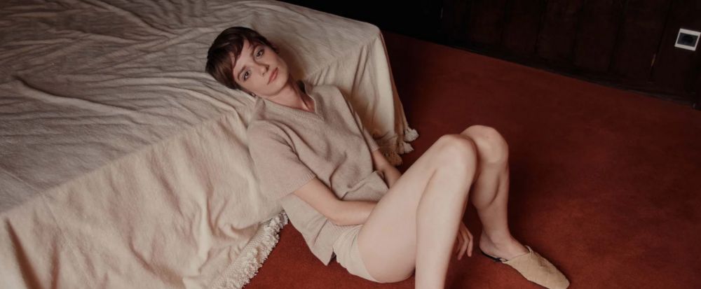 A woman with short brown hair sits on a red carpet, leaning against a cream bed, wearing a beige knit outfit and slippers, looking at the camera.