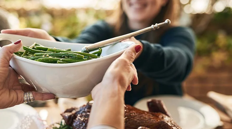 A woman handing a white bowl filled with green beans to another woman over a cooked chicken