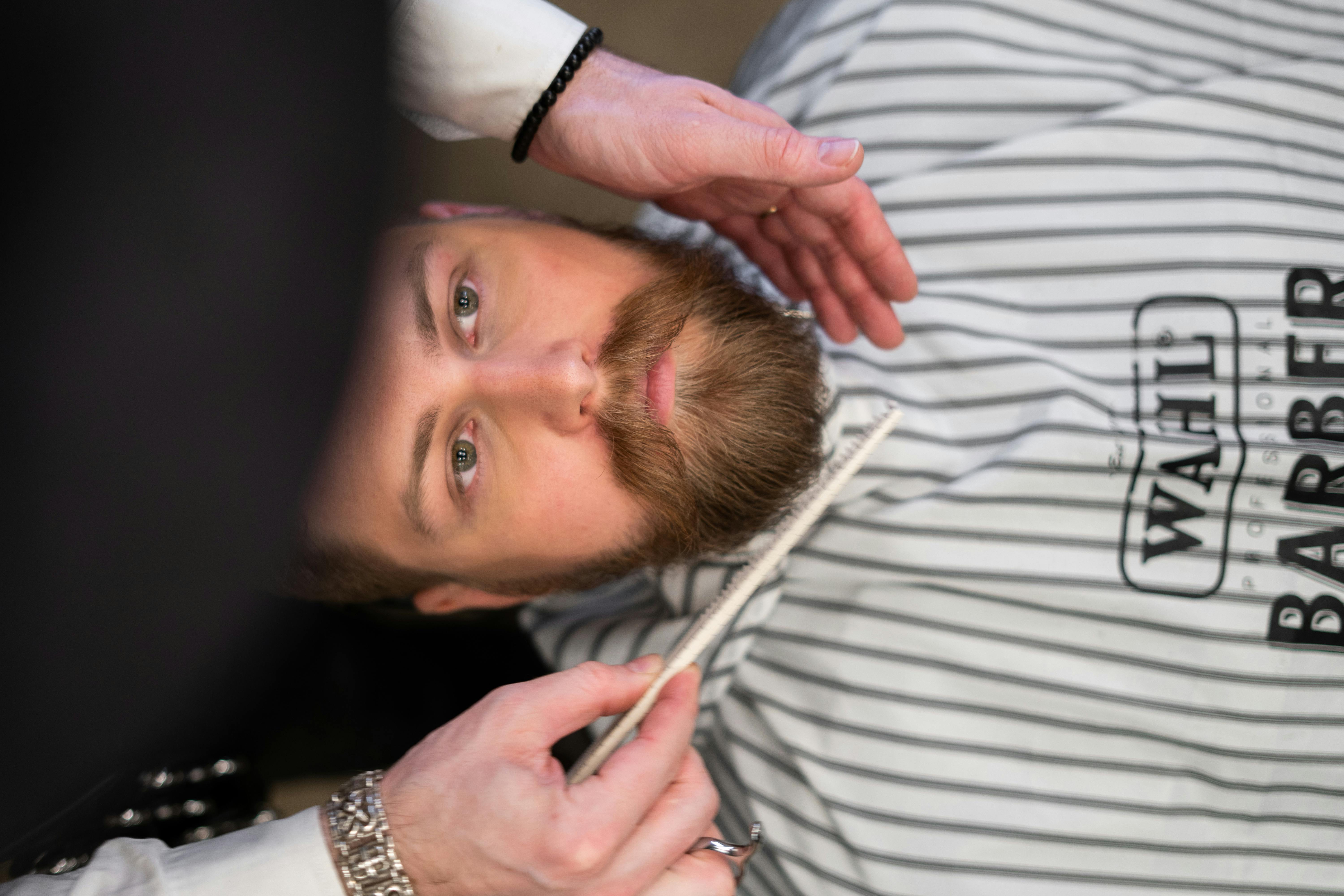 A man’s face facing up and a barber’s hands holding and brushing his beard.