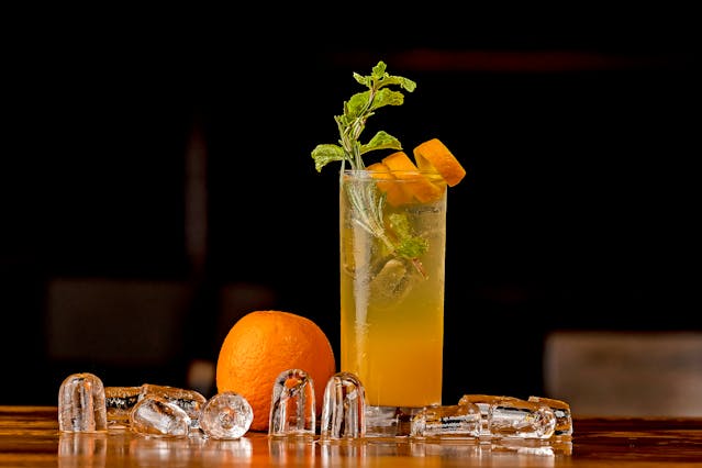 An orange mocktail topped with ice, an orange twist and mint leaves on a tabletop with ice and orange in front of the glass.