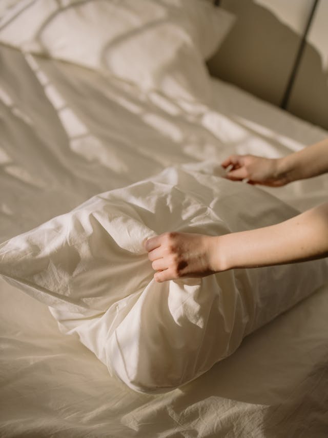 A person fluffing up and adjusting a white pillow while making a white bed.