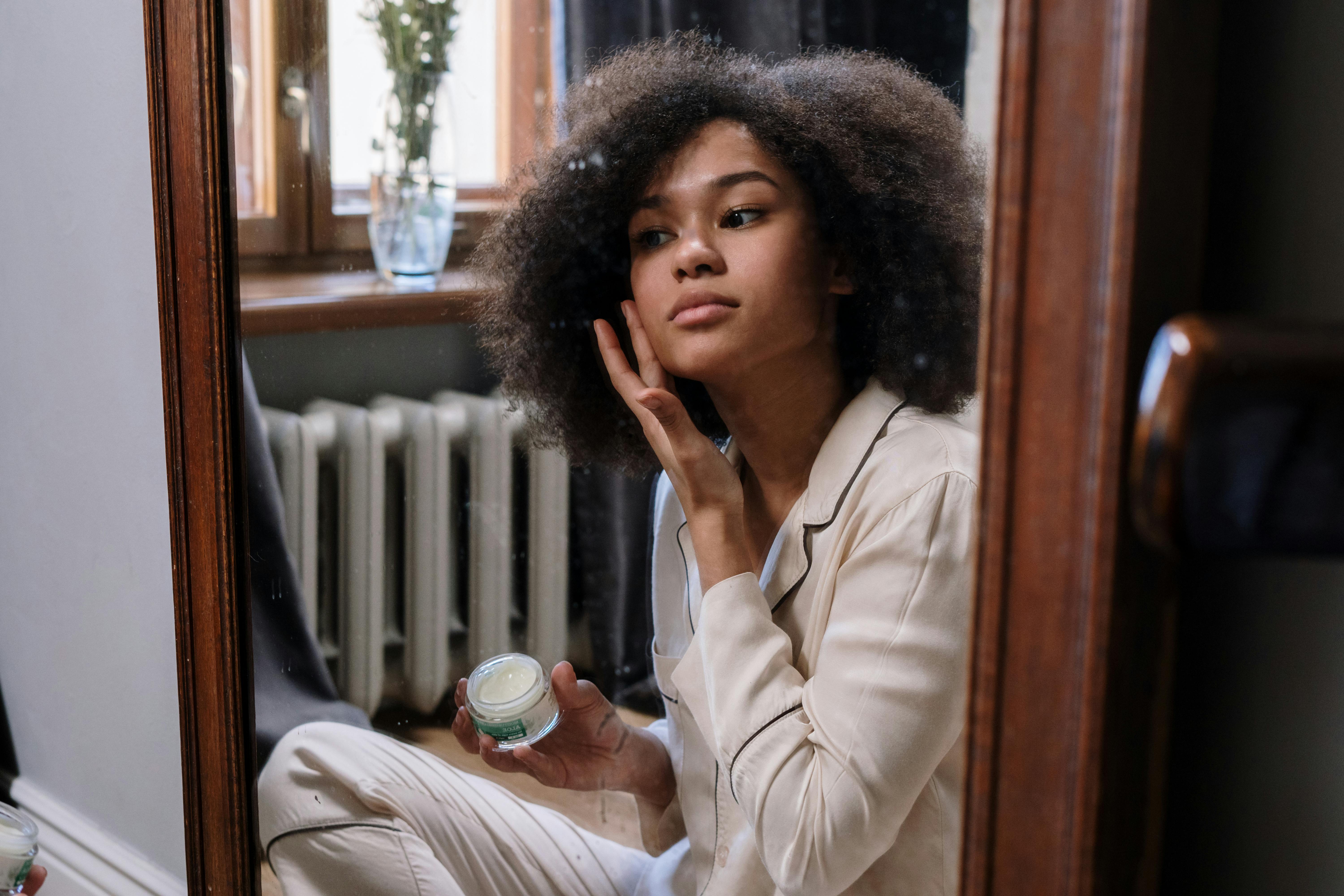 A young lady looking into the mirror and applying a facial cream to her face.