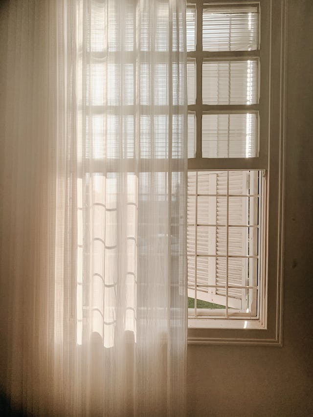 A set of white curtains hung in front of an open window of an apartment in a city.