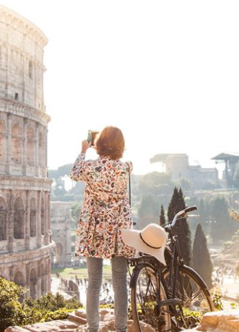 A woman with a black bicycle by her side taking pictures of Rome's  Colosseum using her mobile phone