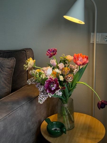 A colourful bouquet of tulips and other flowers in a glass vase on a wooden side table
