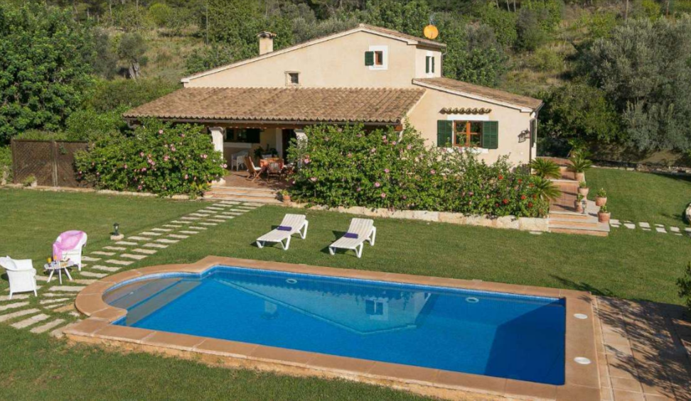 The Ca Na Reieta villa backyard with a pool, white pool chairs, and brown oak patio chairs in a quiet nature surrounding it