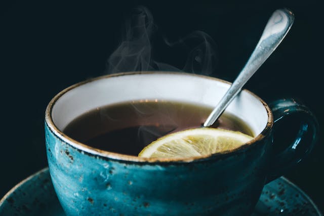 A blue ceramic cup filled with a slice of lemon and a spoon floating in a type of tea.