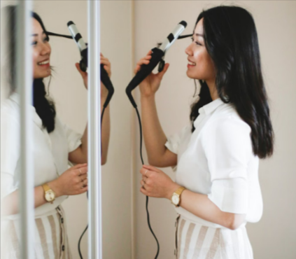 Asian woman using a ghd curling iron in front of a mirror.
