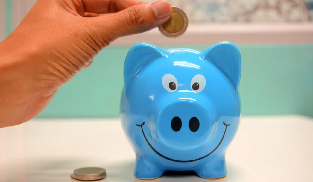 Hand putting a coin in a blue ceramic piggy bank. Background has a painting and is blurred
