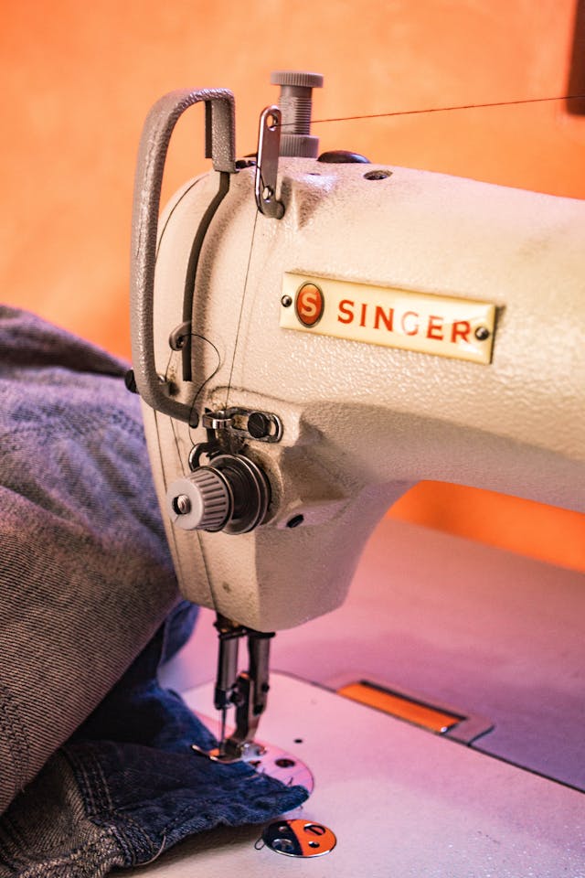 A white and grey Singer sewing machine that is stitching a hem on a grey denim piece of fabric