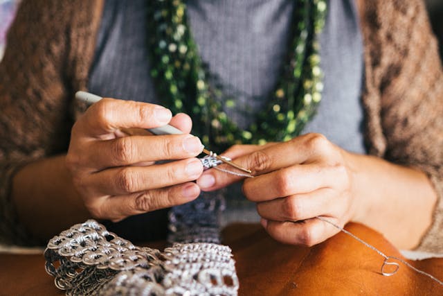 An elderly woman wearing a large green beaded necklace while knitting using a knitting needle and grey yarn.