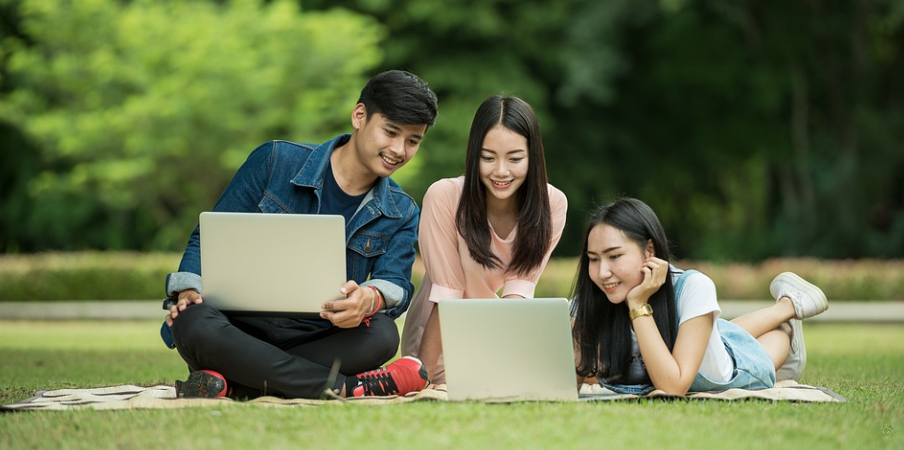 Three students sitting on the grass in the park, searching for OnePlus voucher codes with the laptops