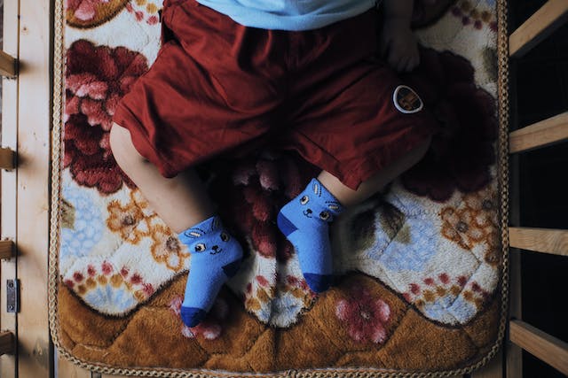 A baby in red pants and blue socks lying on a brown blanket in a cot