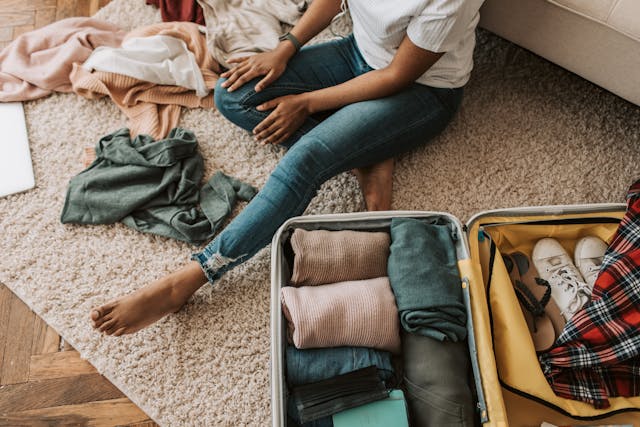 A sitting on a fluffy cream carpet sorting and packing clothes, shoes and other belongings into a suitcase