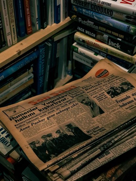 Piled up newspapers with a bookshelves with books in the background