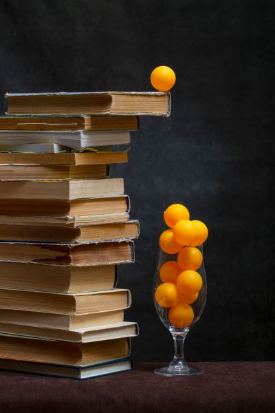 A stack of old books with an orange ball balanced on top and next to a glass full of orange balls