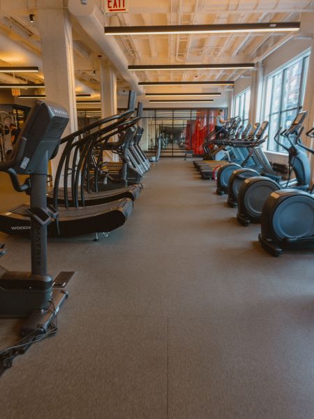 A gym with treadmills and elliptical machines lined up in front of large windows