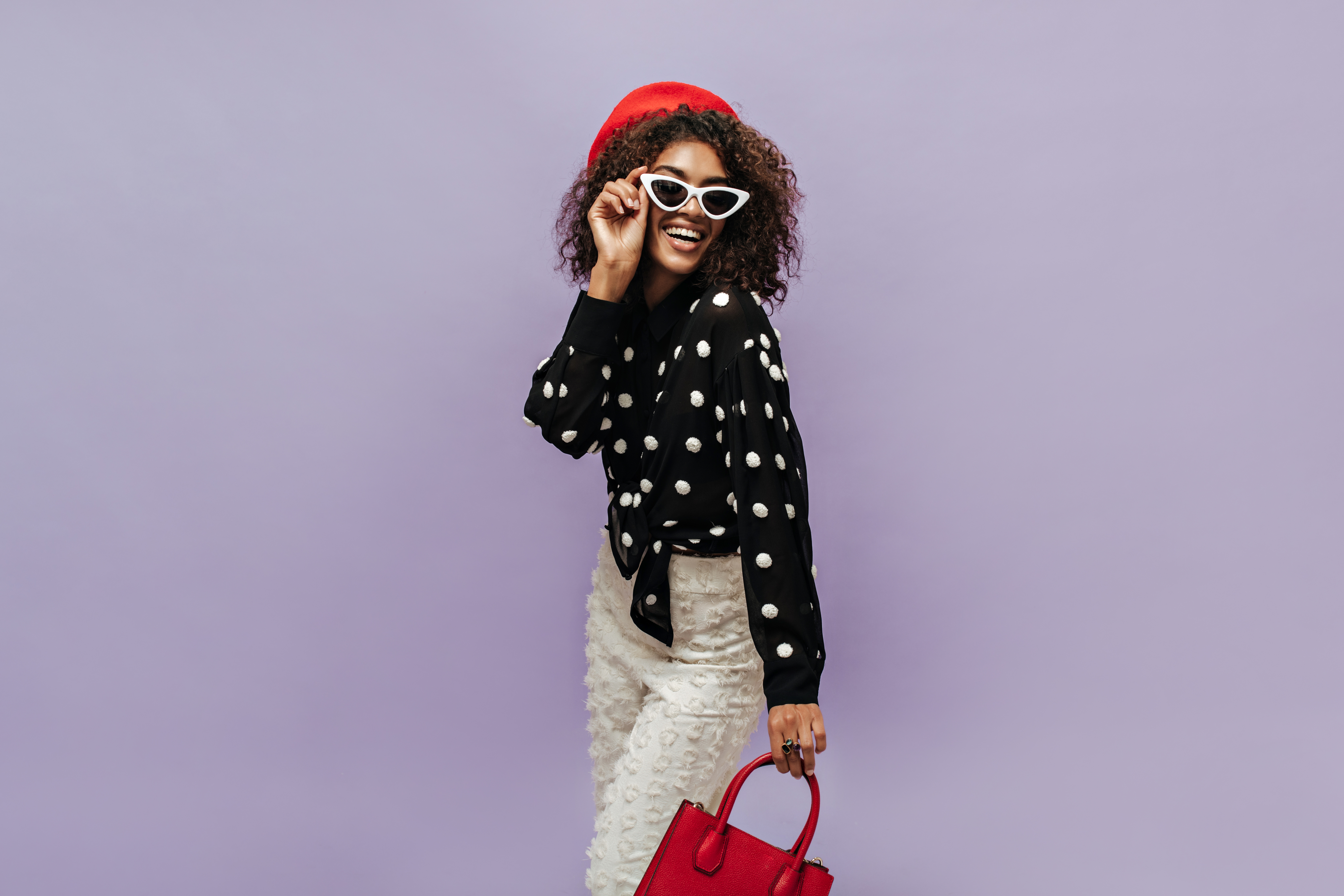 cheerful-brunette-haired-woman-with-red-cap-white-sunglasses-black-shirt-light-pants-smiling-holding-handbag
