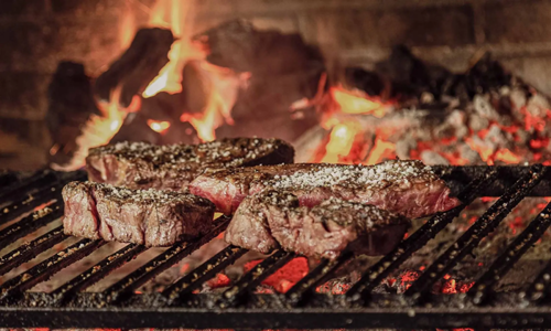 Mehrere Steaks auf einem Grill mit glühender Kohle vor einem Karminfeuer